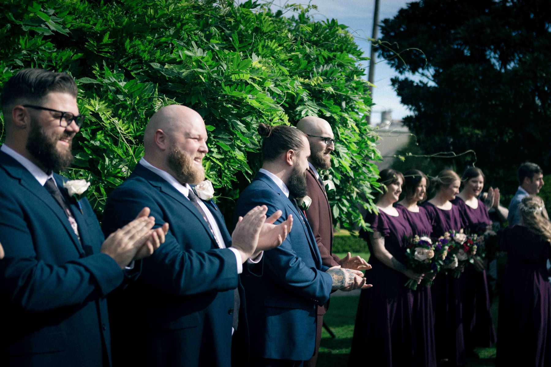 men in tuxedos clapping with women in purple dresses holding boquets of flowers standing nearby