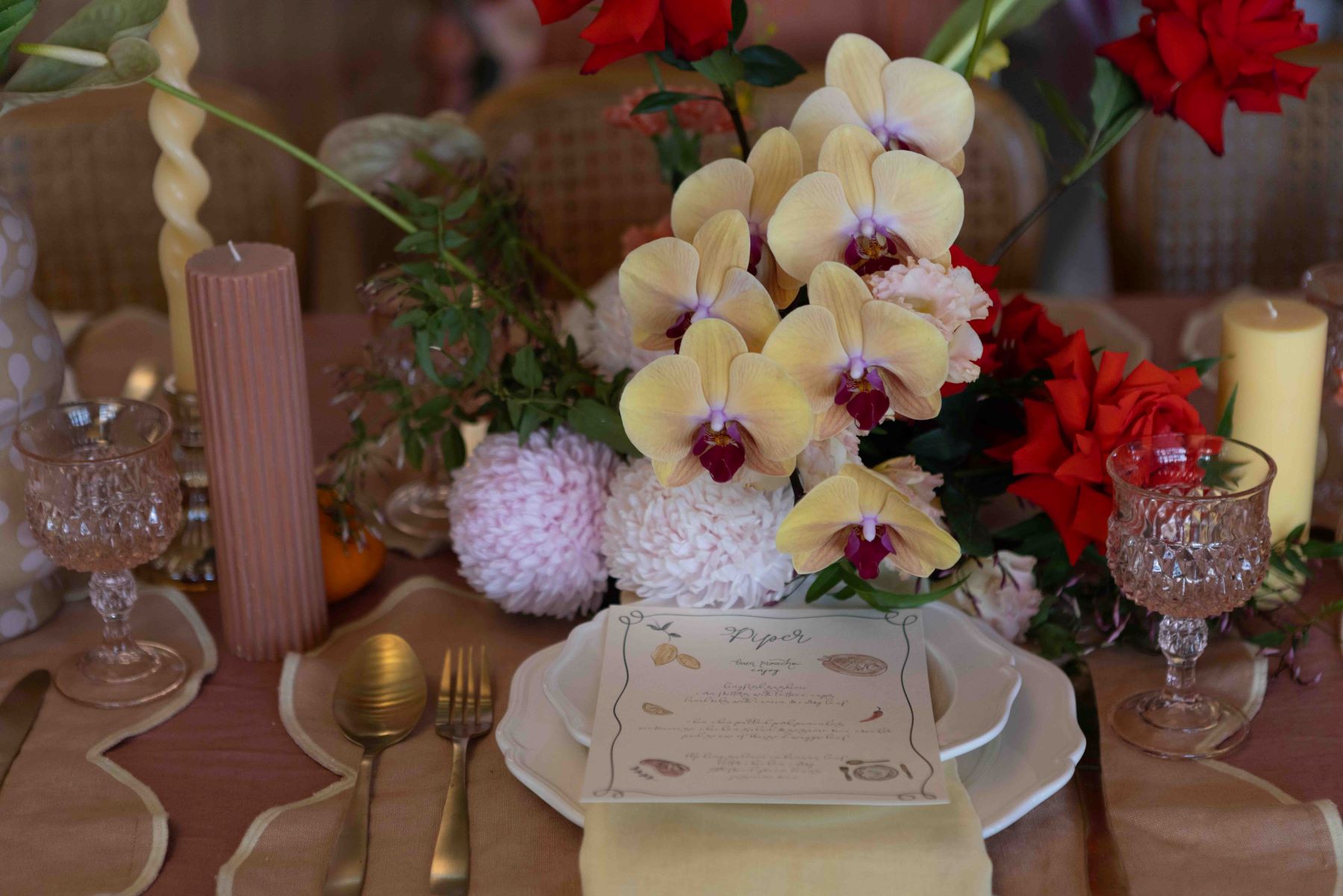 white plates with a wedding invitation and gold silverware, a pink place mat, pink glasses and candles and yellow and maroon flowers all on a table