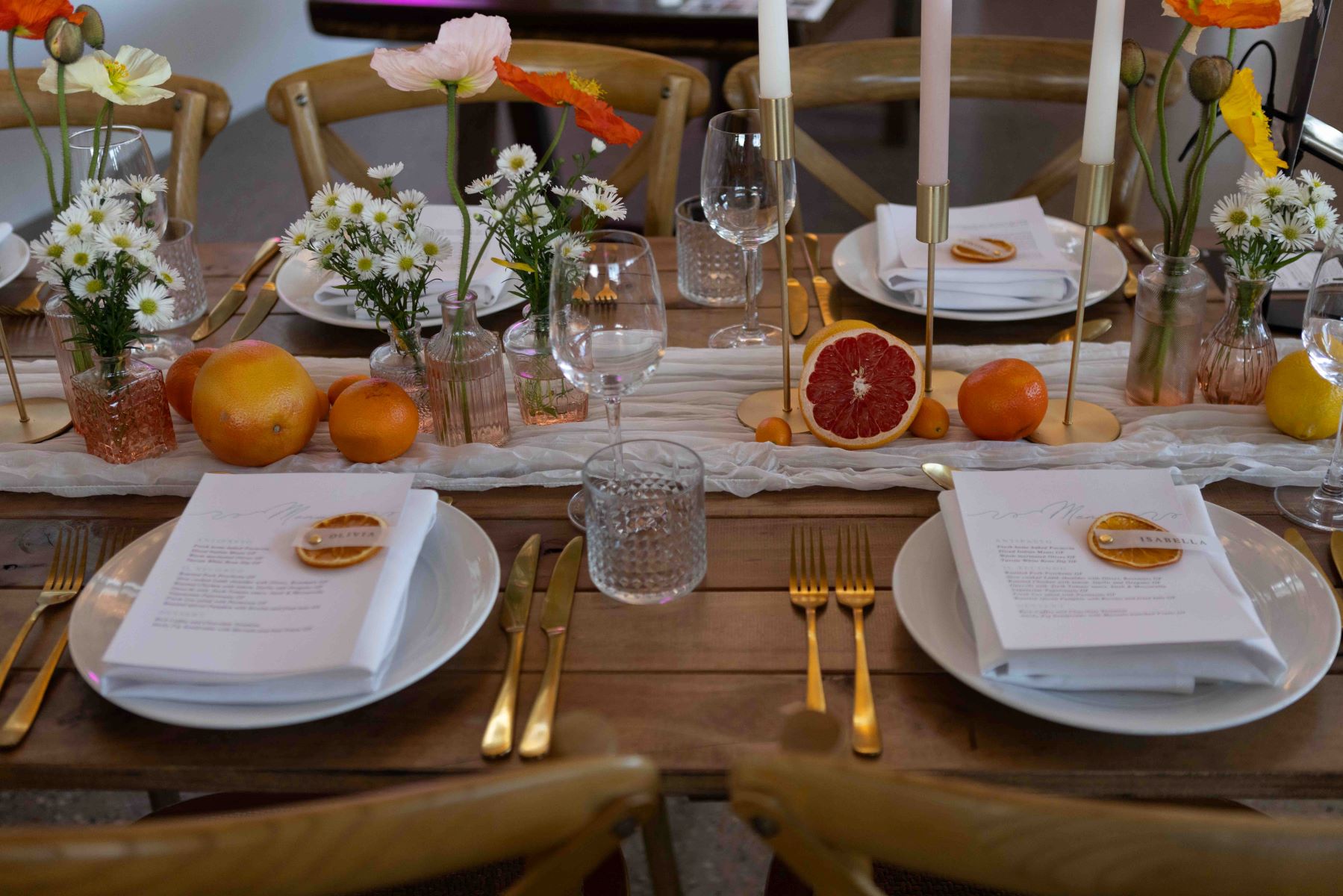 white plates on a wooden table with wedding invitations and gold silverware and glass vases, daisies, fruit, a tablecloth, and candles all on display with wooden chairs surrounding the table