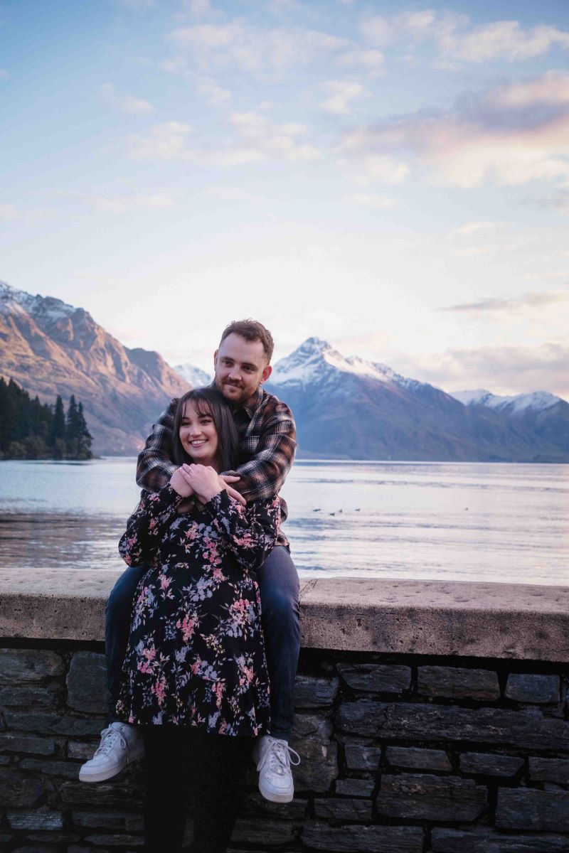 A man and woman next to a lake and surrounded by mountains the man is sitting on a stone ledge and the woman in a floral dress is leaning back into his arms and he has his arms around her shoulders both are smiling the man is wearing a flannel shirt, jeans, and white shoes 