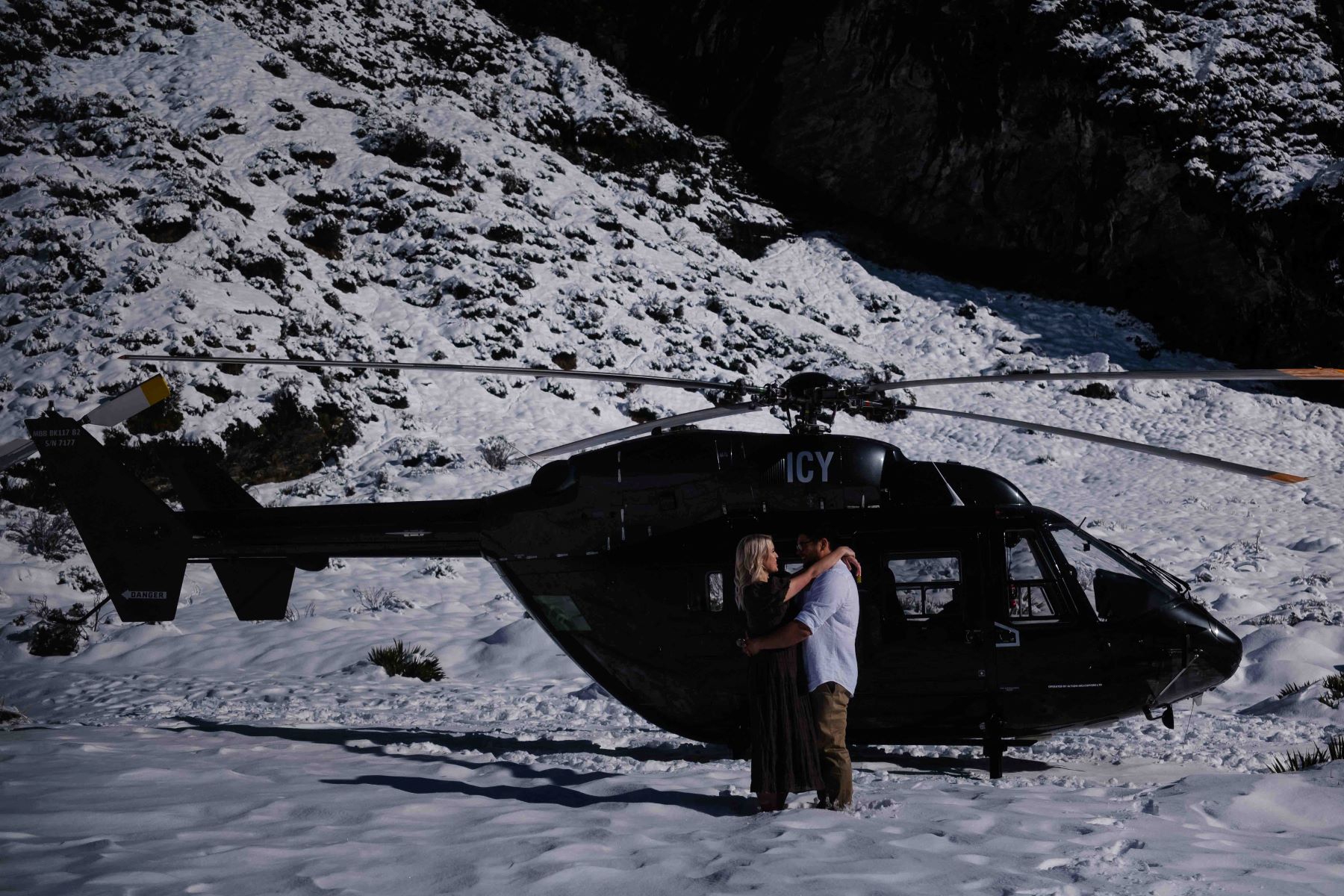 A man and woman are hugging each other in front of a black helicopter on a mountain covered in snow the woman has her arms around the man's shoulders and the man has his arms around her waist the woman is wearing a dress and the man is wearing a button up blue shirt and tan slakc 
