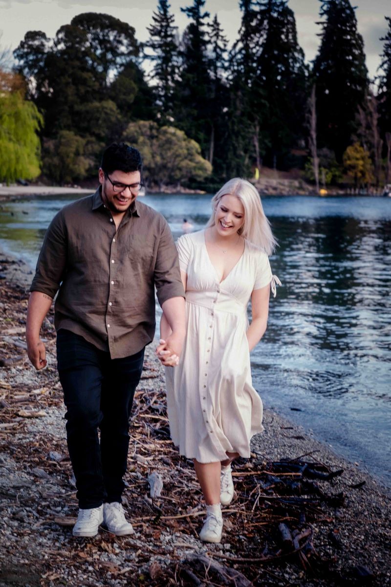 A man and woman are walking next to a lake the woman is wearing a white dress and has one hand in her pocket and is holding the hand of her partner the man is wearing a button up and jeans and white sneakers and glasses both the man and the woman are laughing 