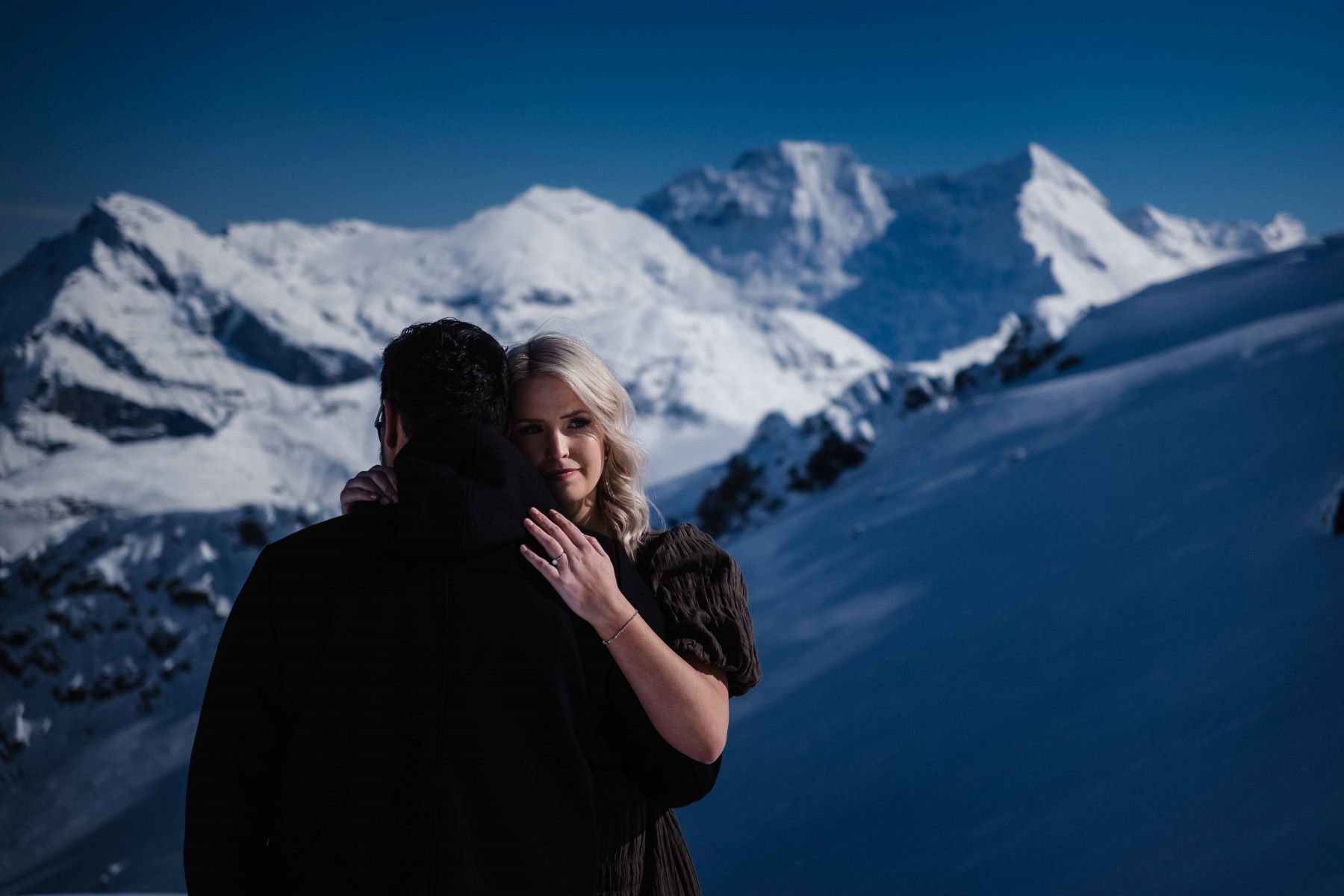 couple hugging each other the woman has her arm on her partner's shoulder and the man's back is facing the mountains they are surrounded by snow and snow-capped mountains
