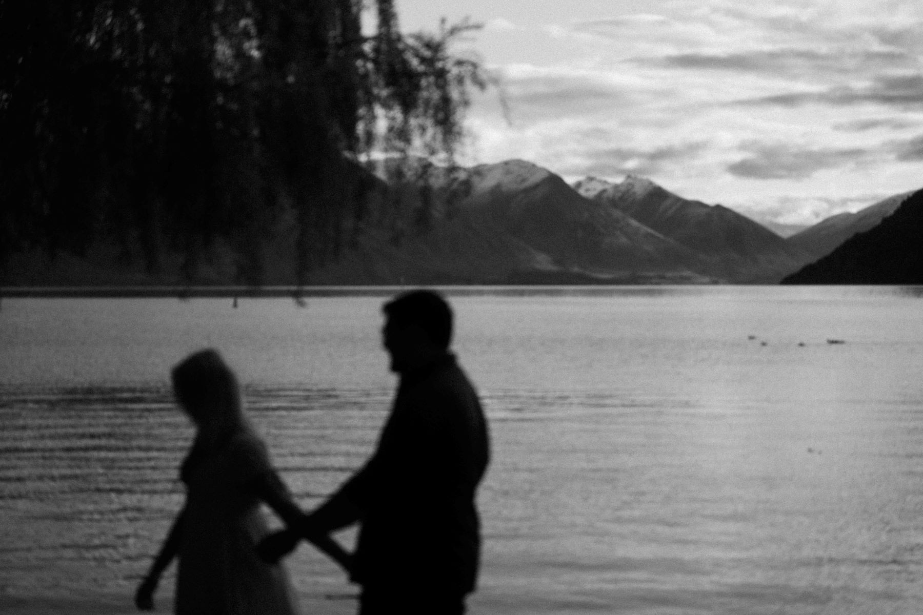 A silhouette image in black and white of a man and woman holding hands and walking next to a lake and surrounded by mountains 