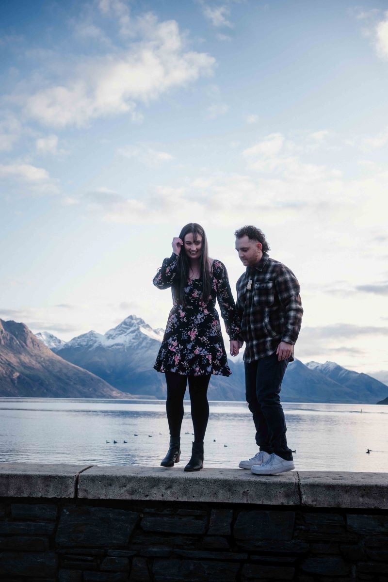 A man and woman holding hands and standing on a ledge in front of a lake with mountains in the background the woman has on a floral dress and tights and black boots and the man has on a flannel, jeans, and white shoes and is looking towards the ground the woman is also smiling and looking towards the ground 