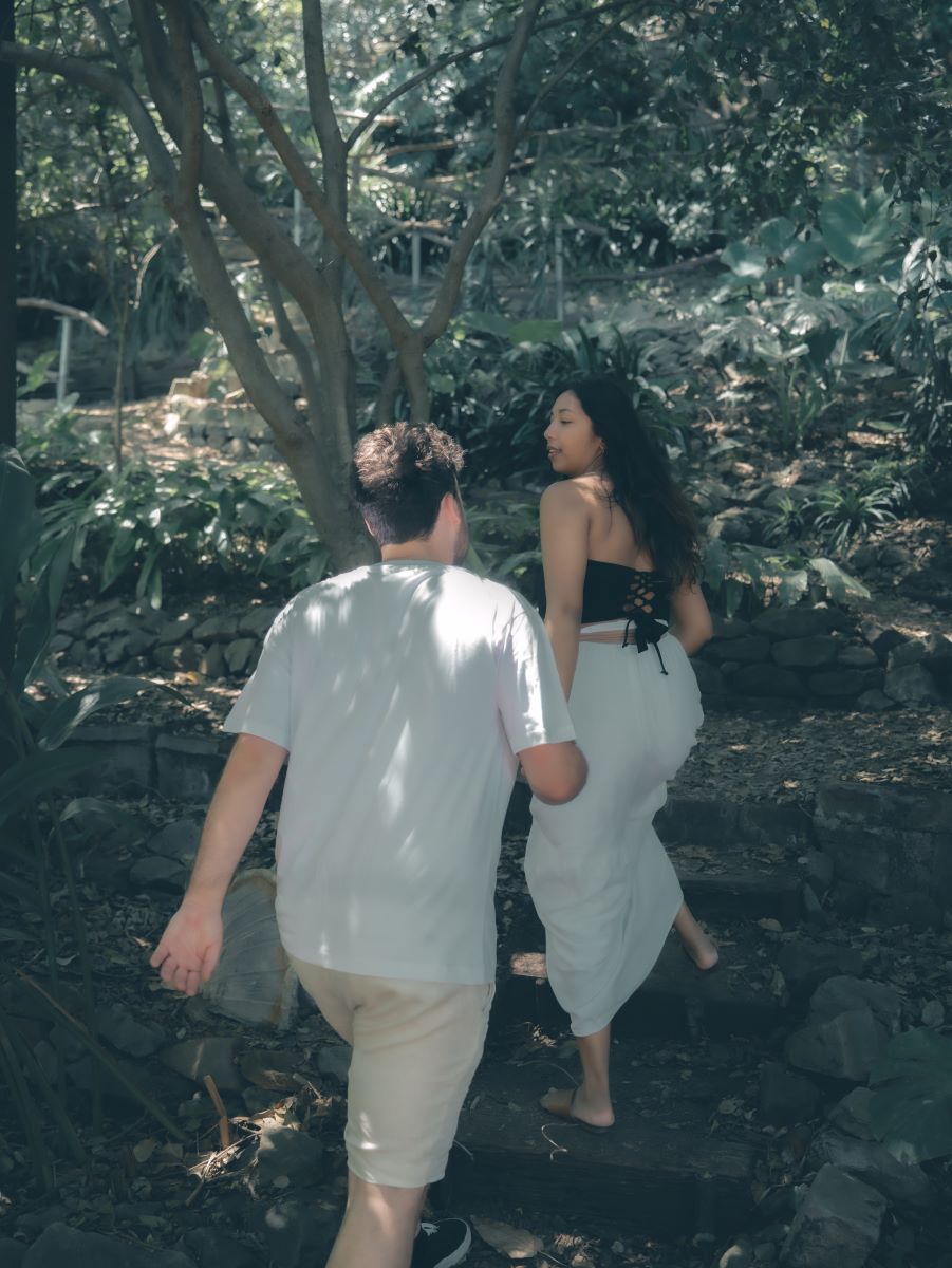 A couple walking through a garden together the woman is weraing a black bandana top and white skirt and the man is wearing a white shirt and tan pants they are both holding hands 
