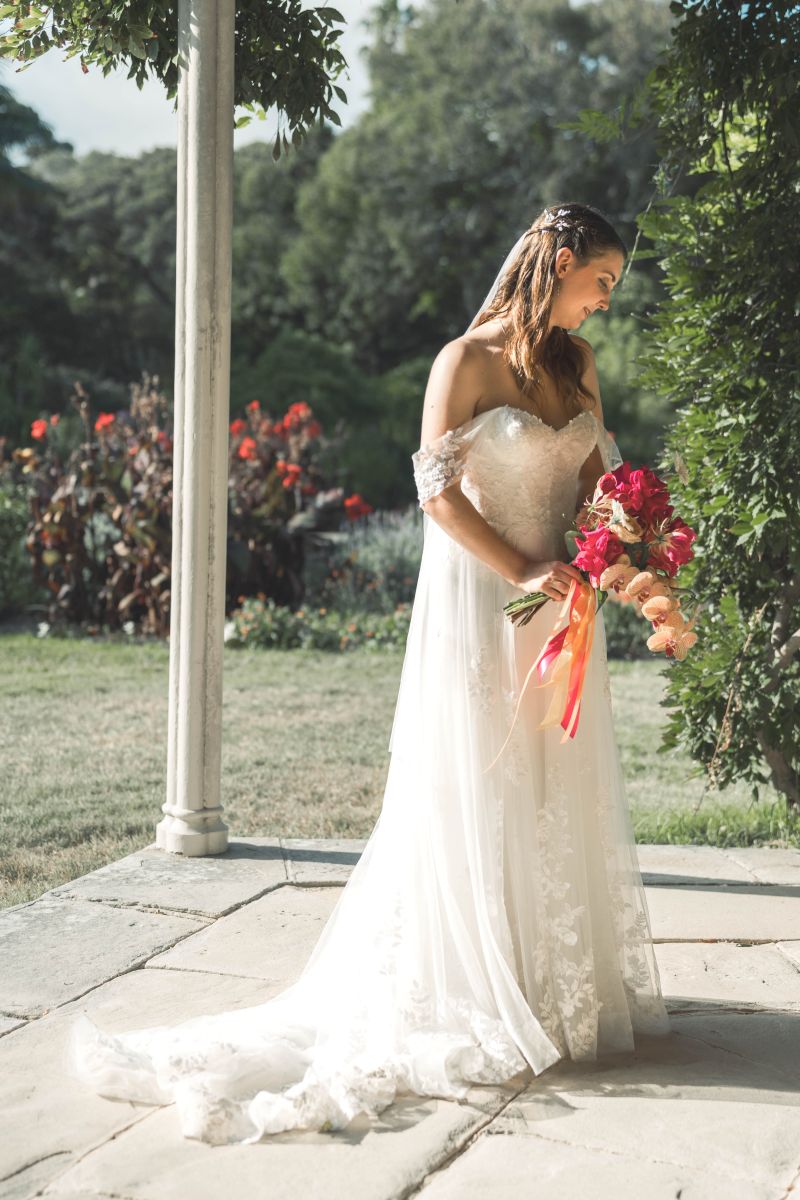 Bride posing thoughtfully in white lace dress with red bouquet in the gardens at Vaucluse estate house—Sydney wedding photographer specializing in authentic bridal portraits