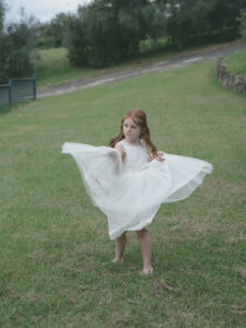 Young flower girl in a white dress twirling joyfully on the lawn during a wedding celebration—candid wedding photographer Sydney documenting unguarded childhood joy and authentic wedding day moments